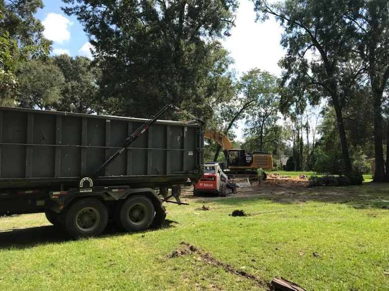 Dump truck, excavator, and skid steer on a grassy area; tree removal in progress.