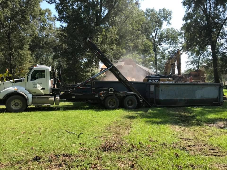 Truck with container lifting debris on a grassy field; in the background, a building.