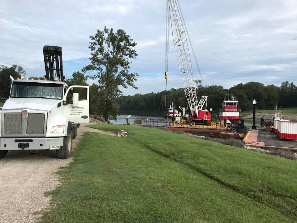 White truck, crane, barge, and tugboat on riverbank with grass and trees.