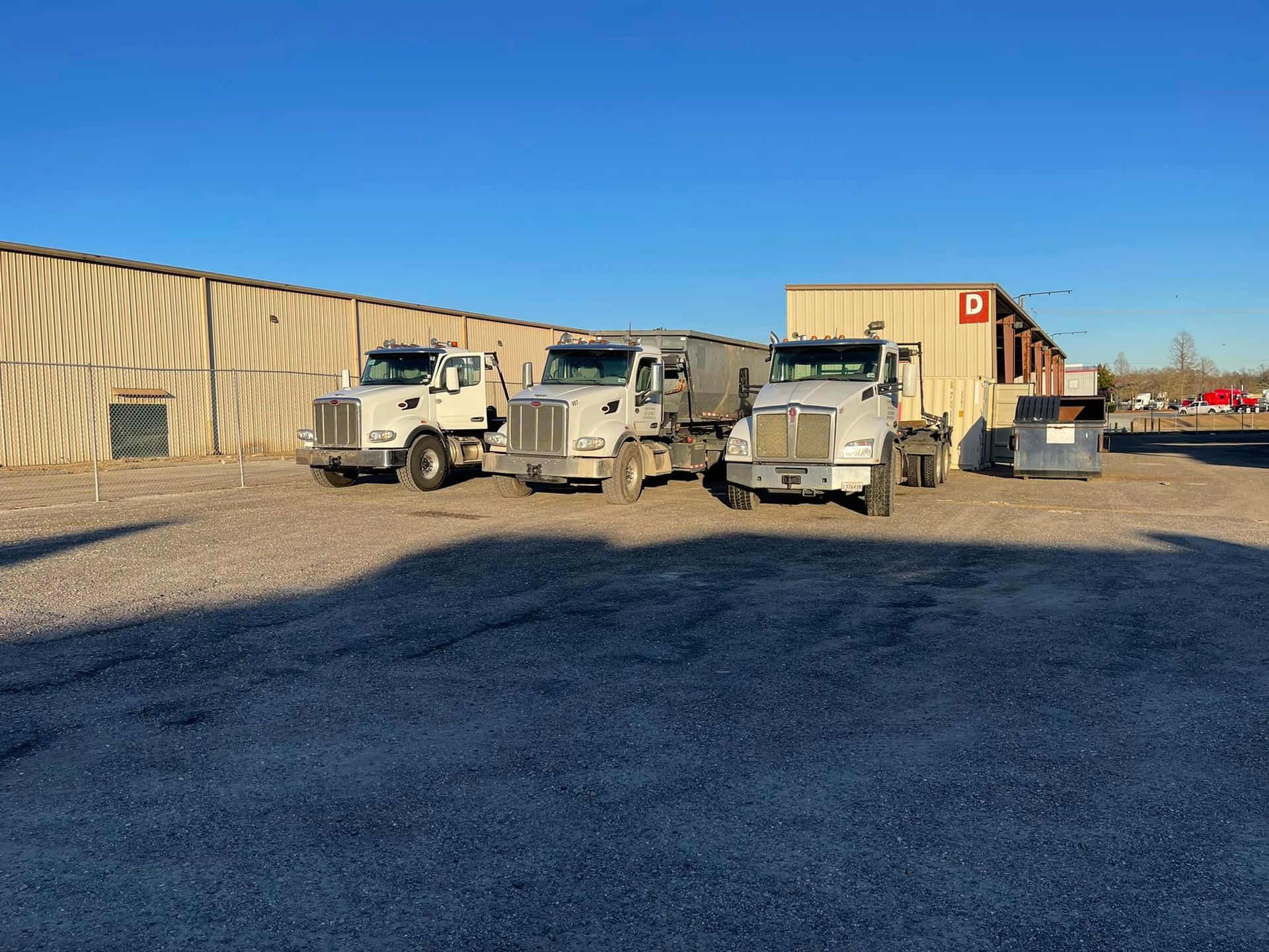 Three white trucks parked in a gravel lot near a tan building on a sunny day.