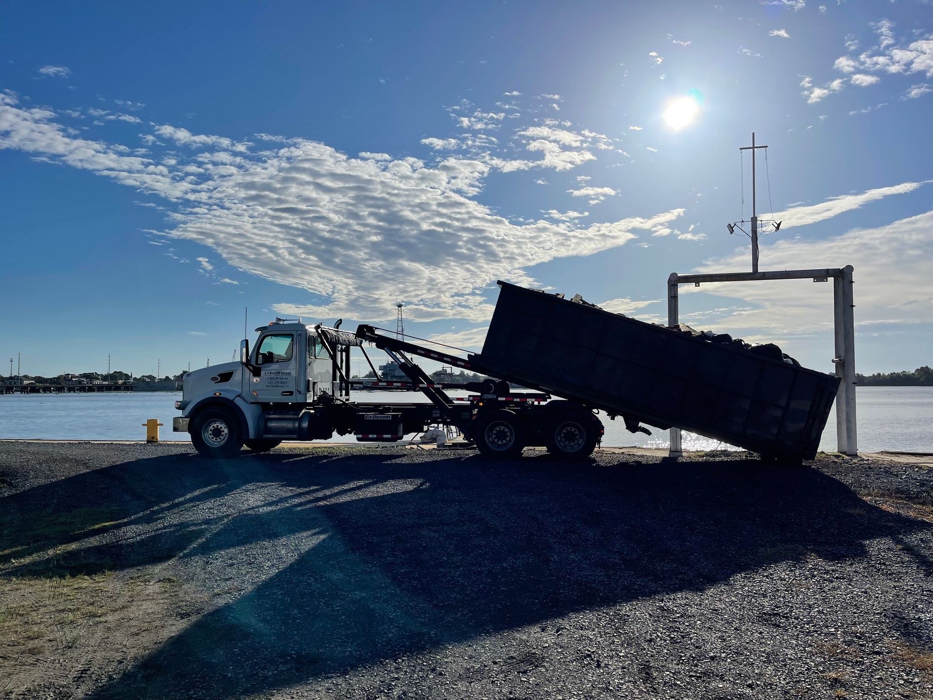 Dump truck unloading waste near water with a bright sun overhead.