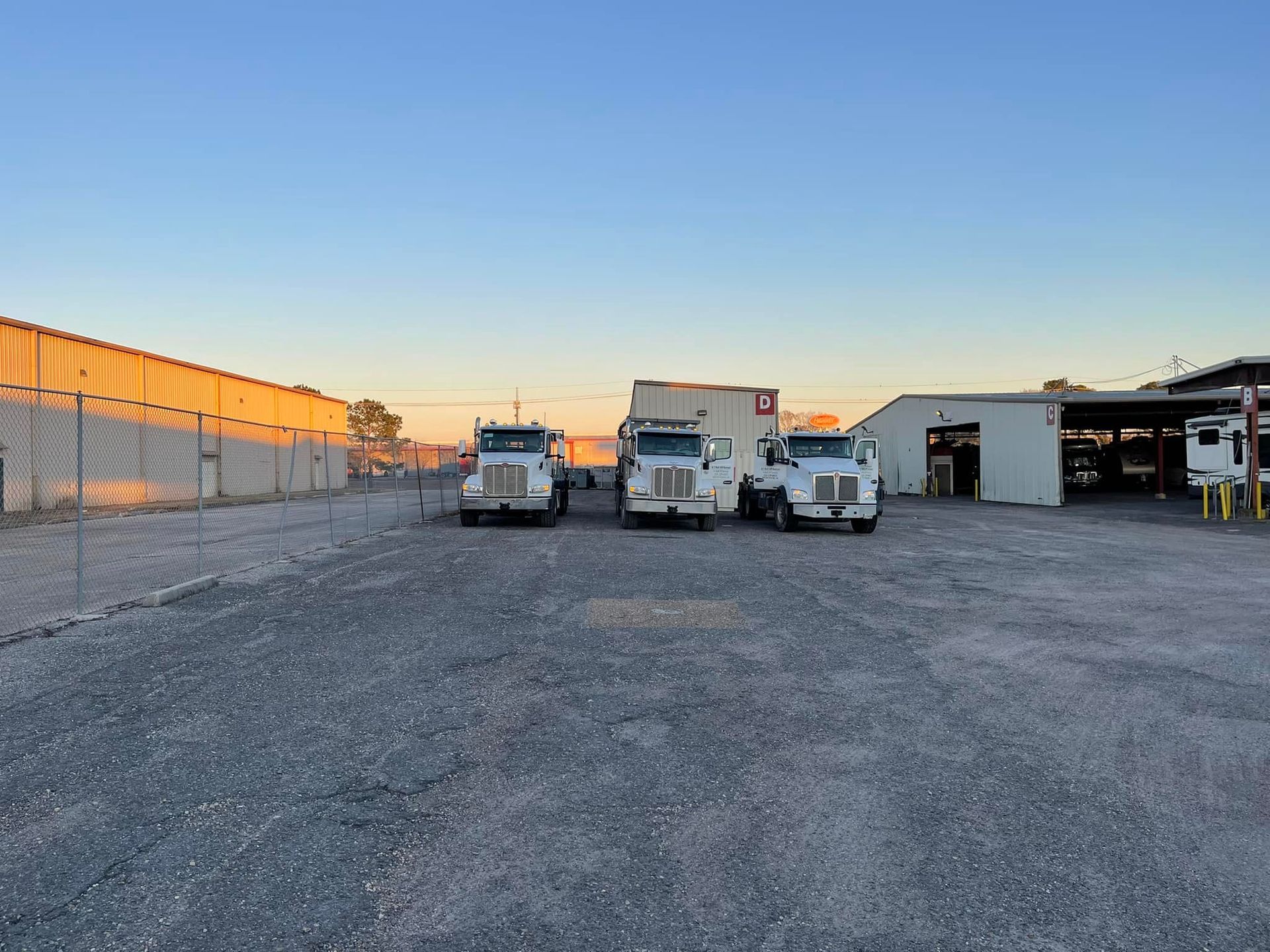 Three white semi-trucks parked on a gravel lot near a fenced area and building at sunset.