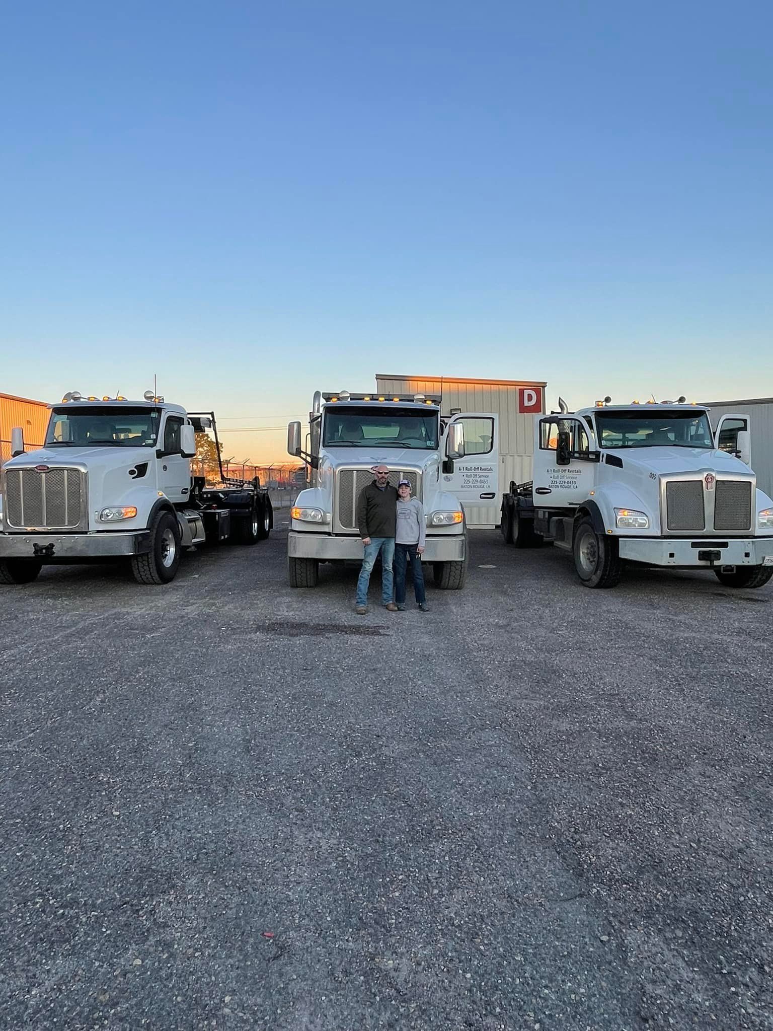 Three white semi-trucks with people standing in front on a gravel lot at dusk.