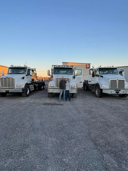 Three white semi-trucks with two people standing between the middle truck, blue sky backdrop.