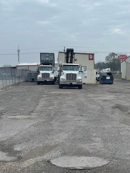 Two white semi trucks with containers on gravel lot in front of industrial buildings.