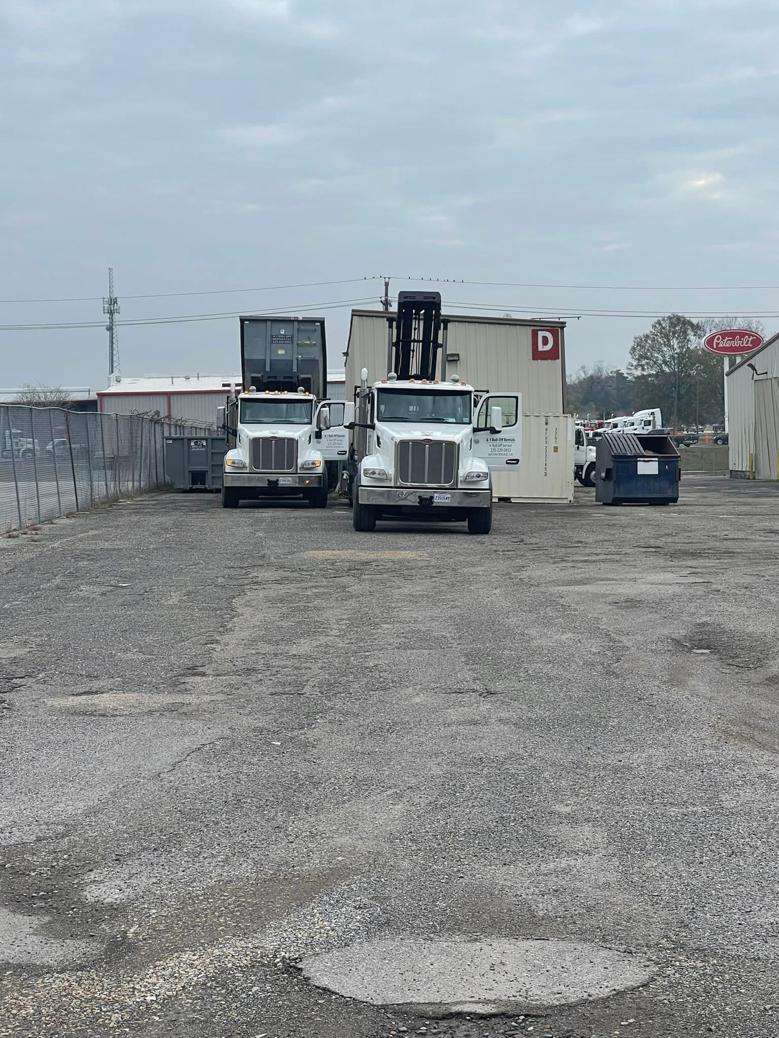 Two white semi-trucks parked on gravel, with a building in the background. Cloudy sky.