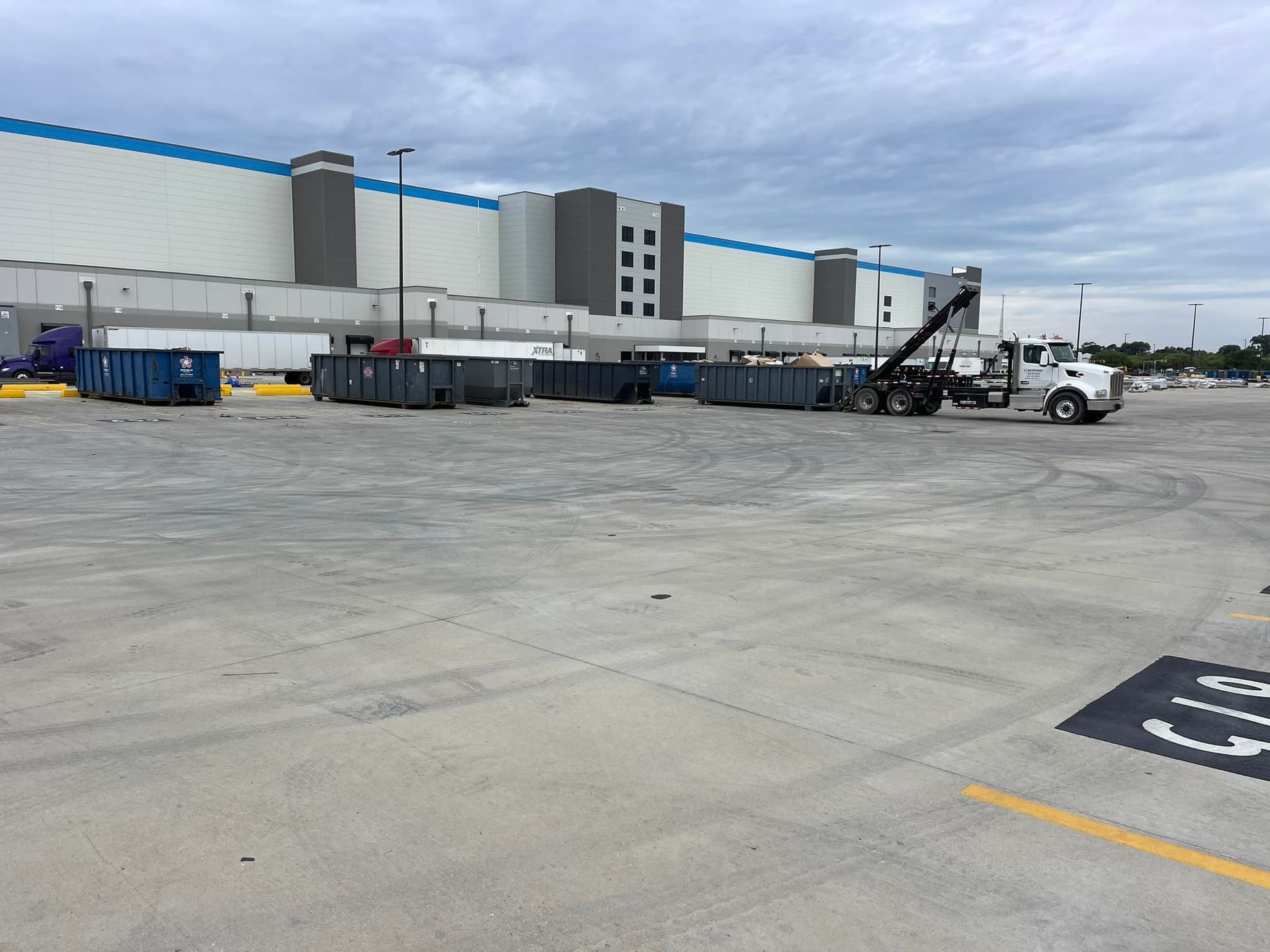 Large parking lot with dumpsters and a crane truck near a large, white warehouse with blue trim. Cloudy sky.