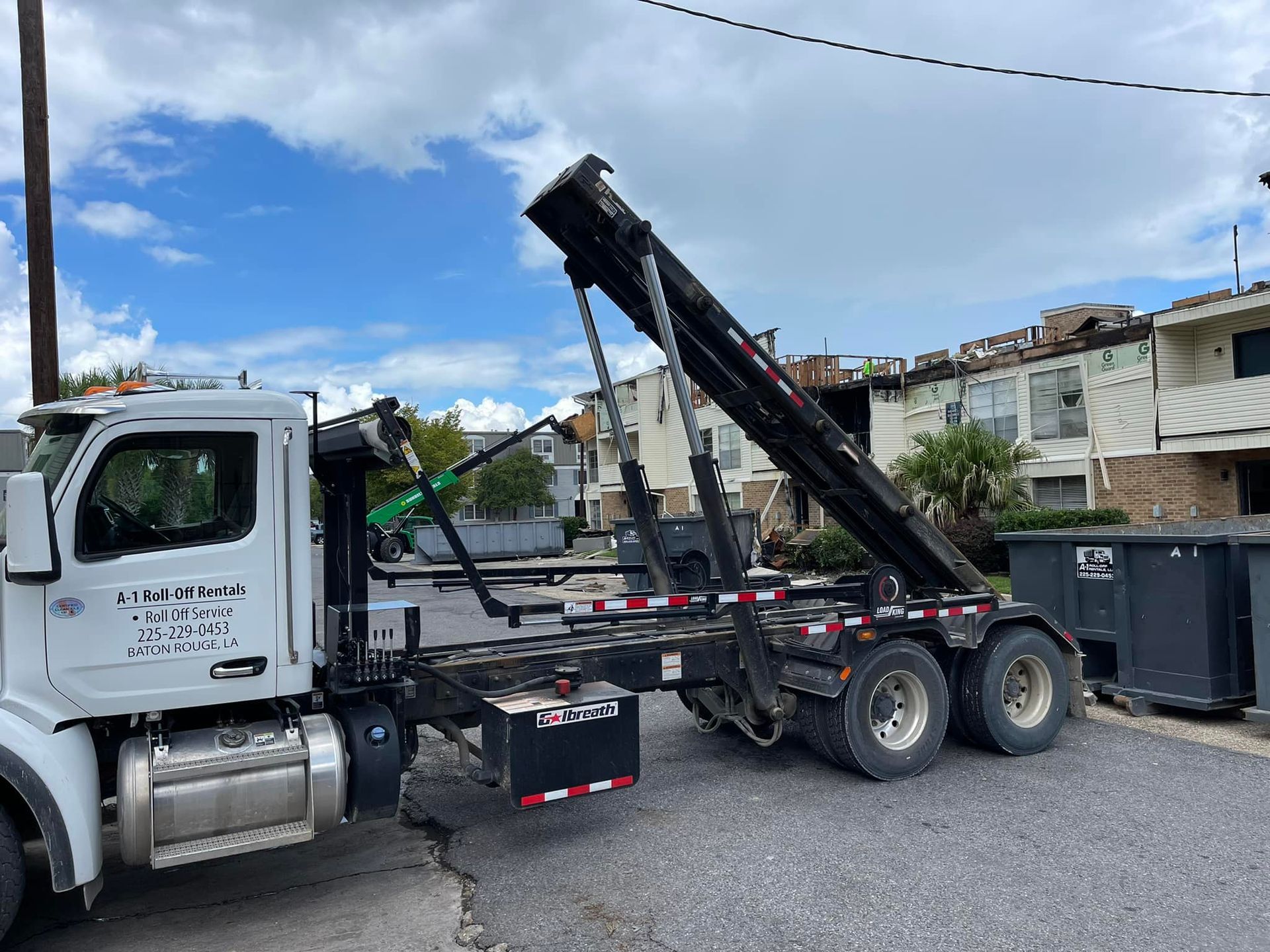 White truck with a dumpster lifting mechanism, parked near a building. Cloudy sky.