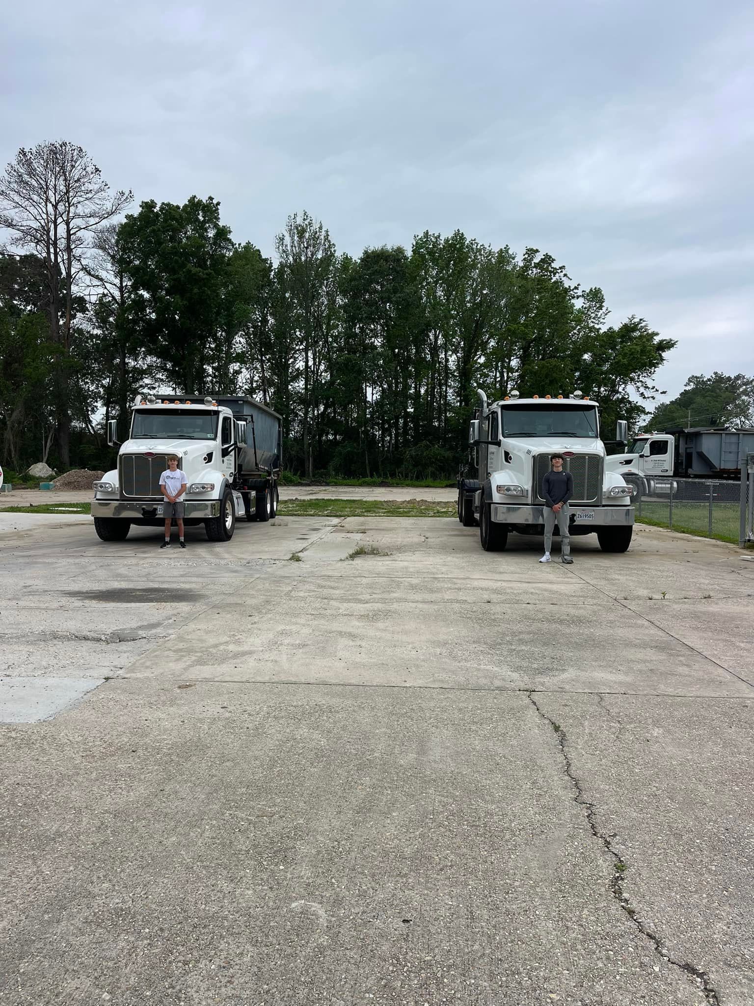 Two white semi-trucks parked on concrete, two people stand between them. Cloudy sky, trees in the background.