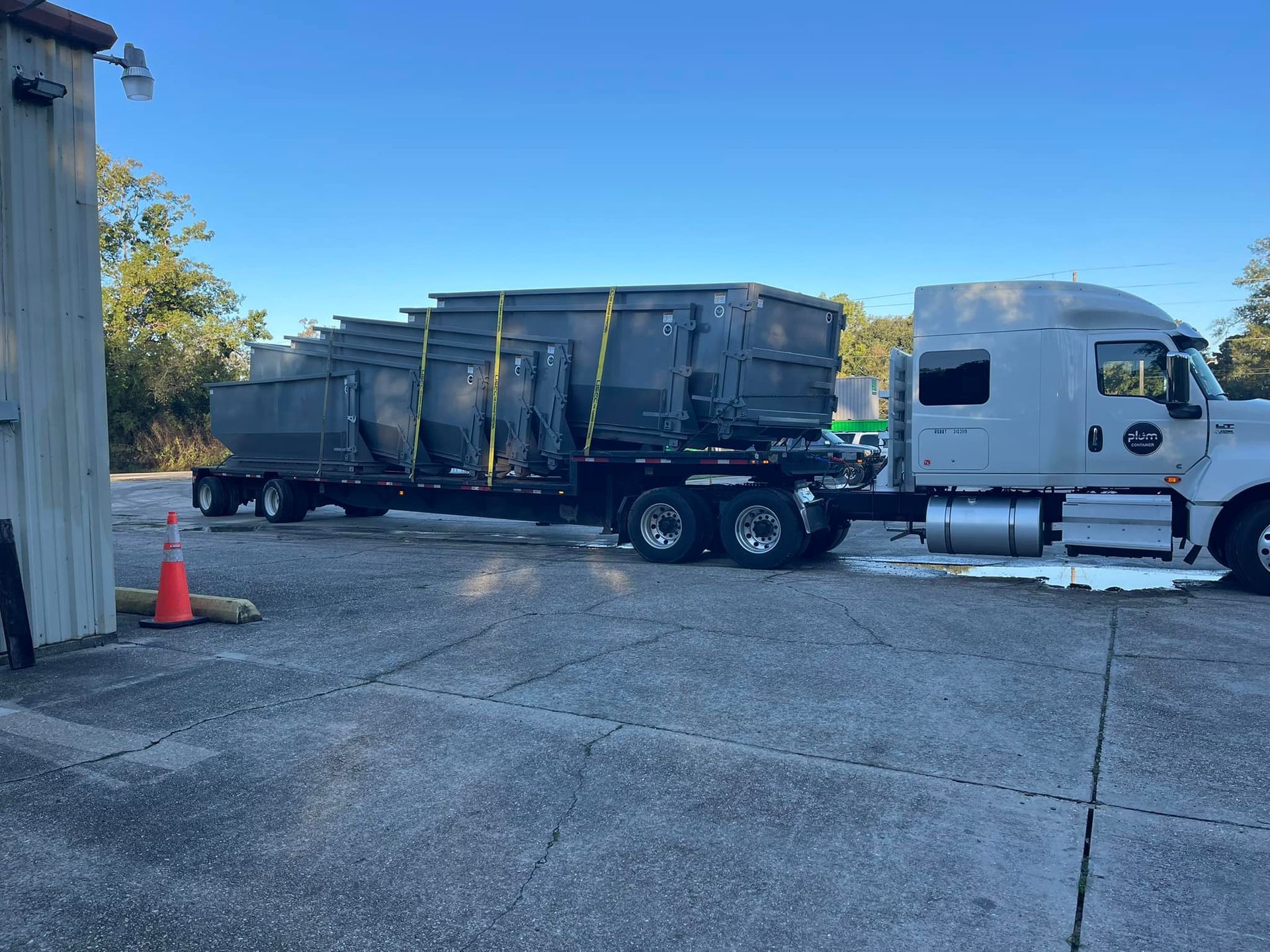 Gray semi-truck with trailer hauling multiple large gray dumpsters on a paved lot.