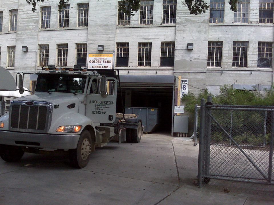 Truck at a loading dock of a multi-story, white building; blue dumpster inside.