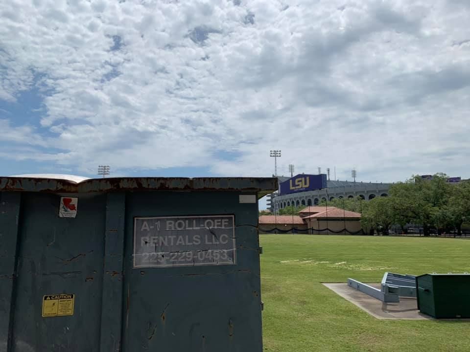 Dumpster in front of a green field with the LSU stadium in the background under a cloudy sky.