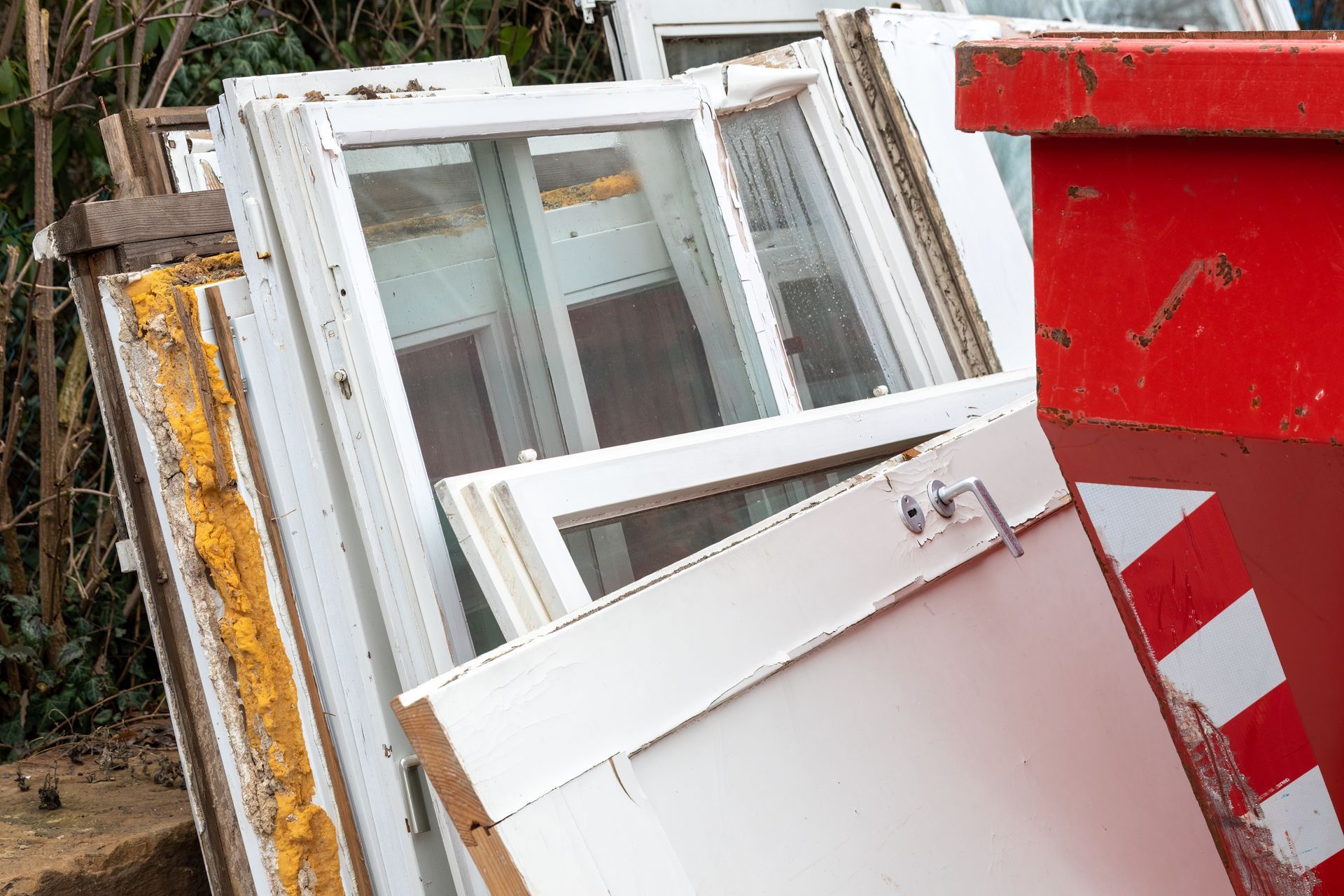 Stacked white window frames and doors beside a red dumpster outdoors.