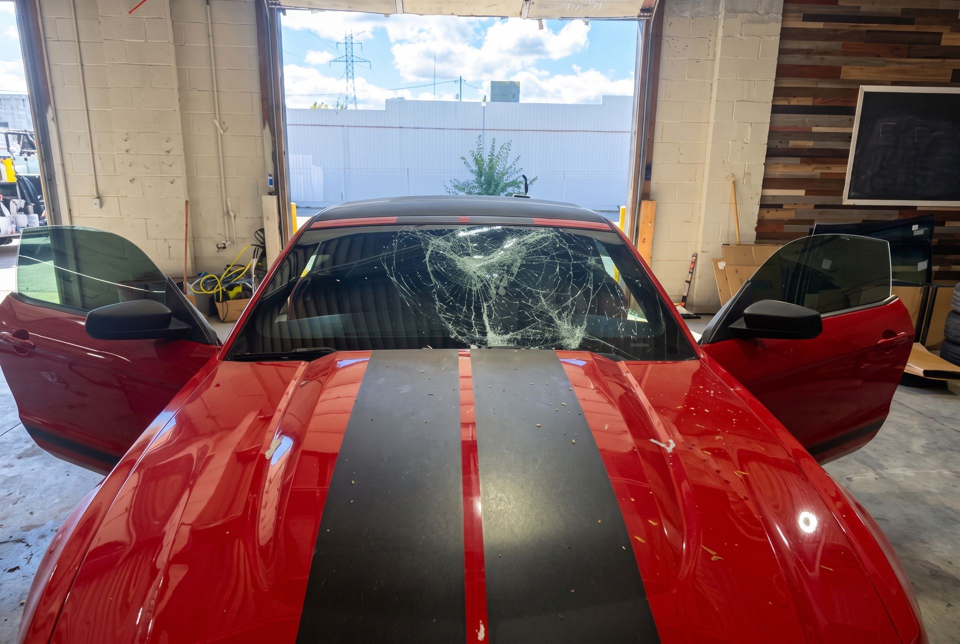 Red sports car with black racing stripes, both doors open in a garage, windshield cracked