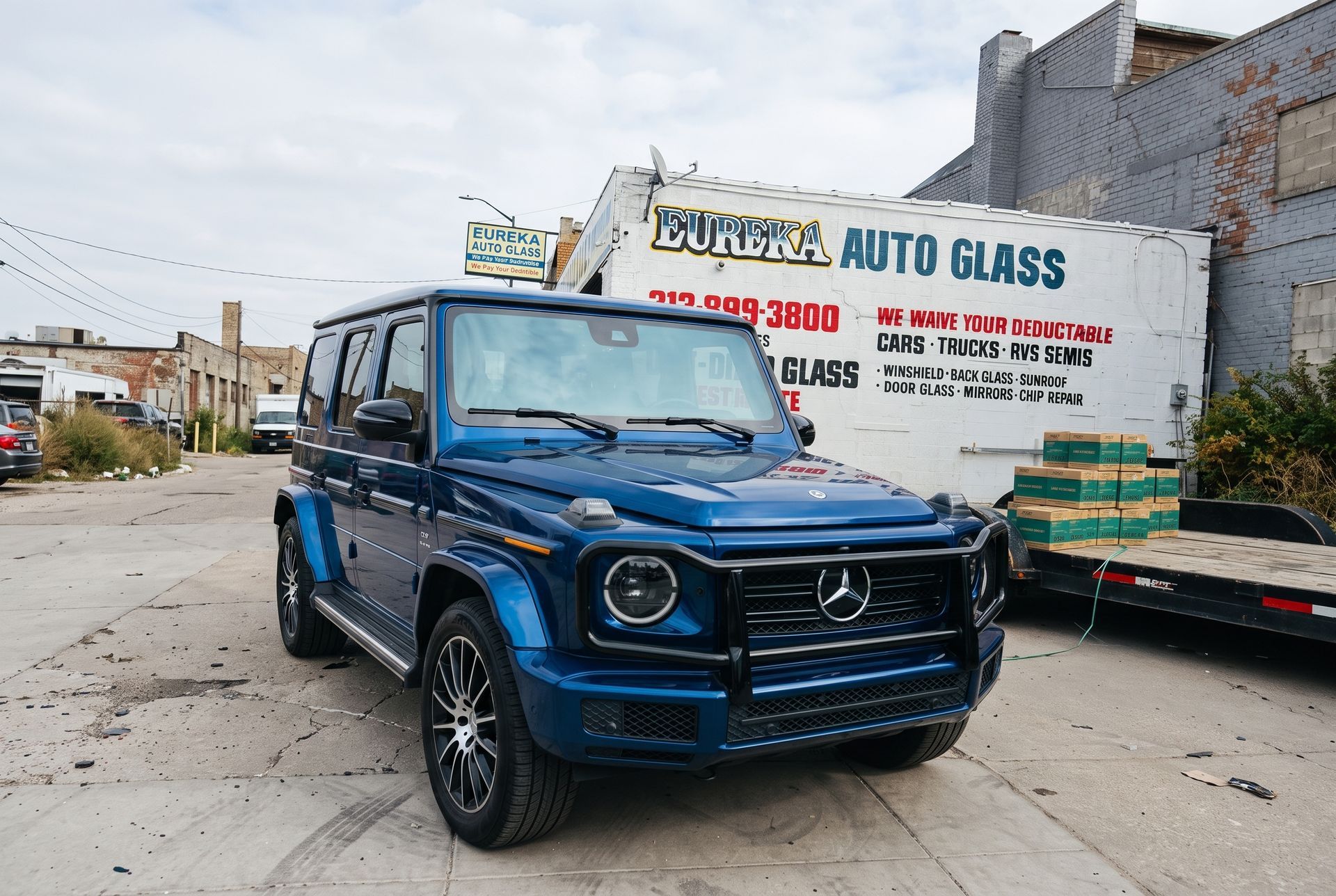 Blue Mercedes G-Class parked outside an auto glass shop on a city street.