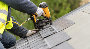 Roofing worker using a nail gun to install gray shingles on a sloped roof