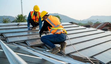Two roof workers in hard hats and orange vests installing metal roofing panels on a steep roof