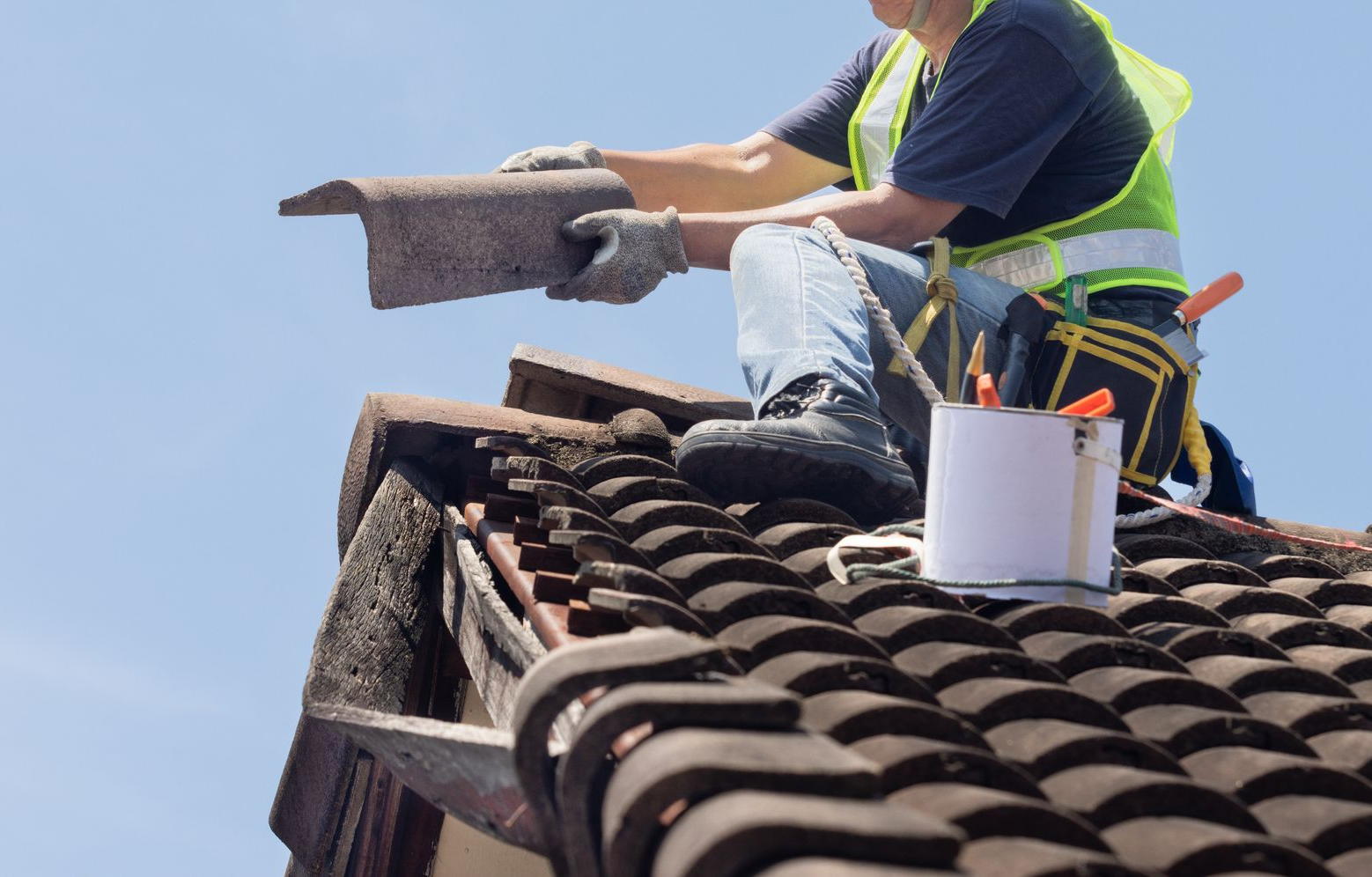 Roofer installing tiles on a house roof with tools and a bucket under a clear blue sky