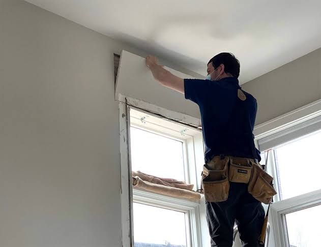 Worker plastering wall above a window in a bright room, wearing a tool belt
