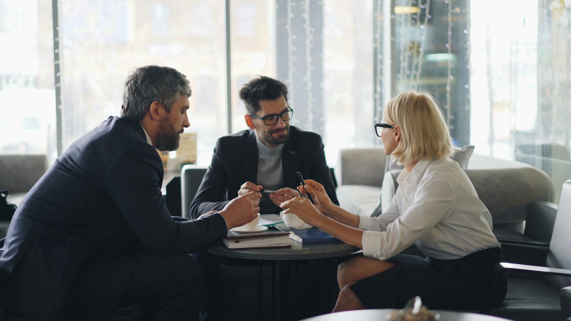 Three people in business attire at a table in a well-lit office, engaged in a discussion, pens in hand.