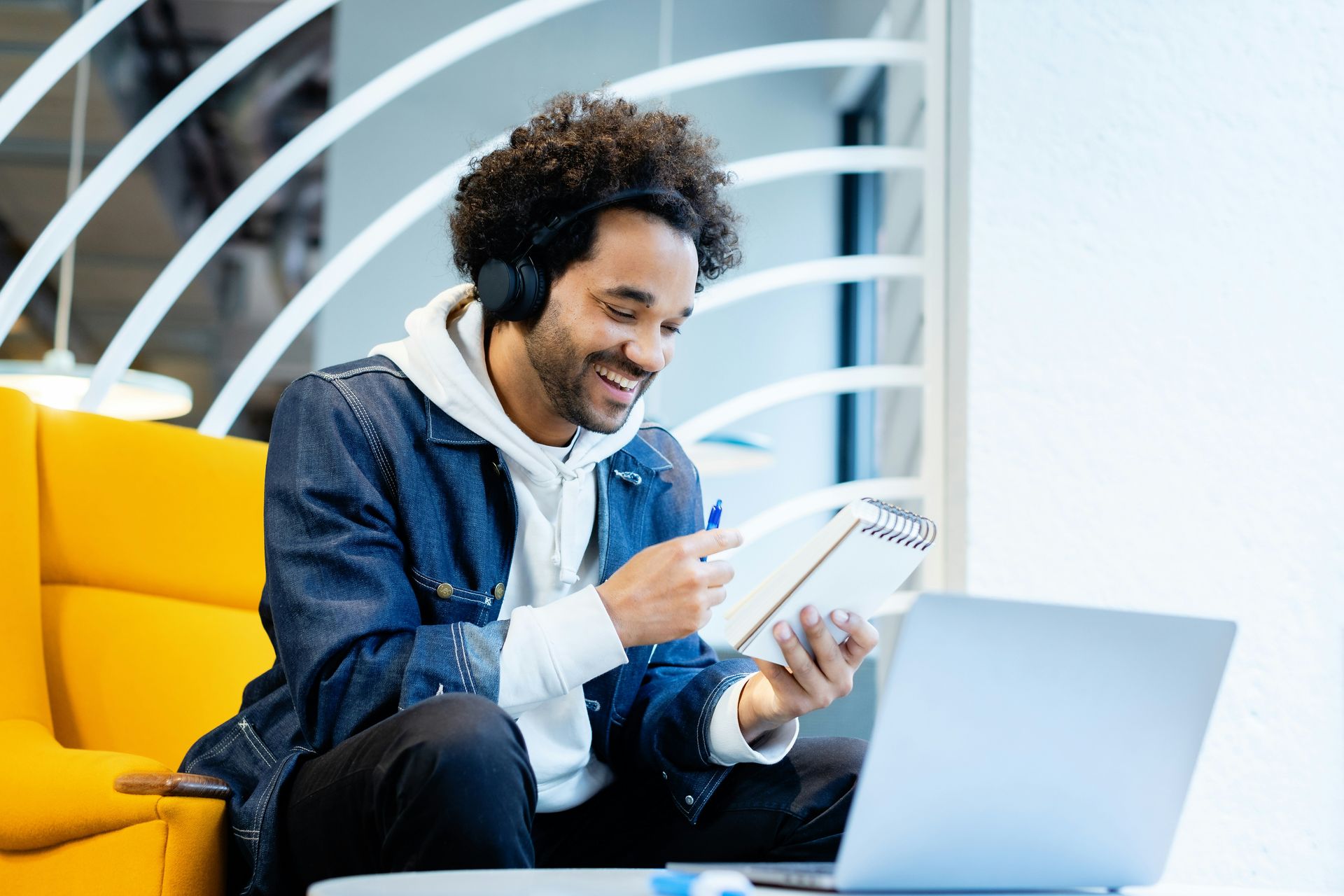 Man wearing headphones, smiling, looking at notebook and laptop in a yellow chair.