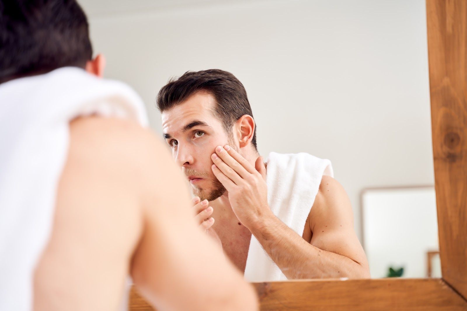 Man in bathroom towel checking his face in the mirror and touching his cheek