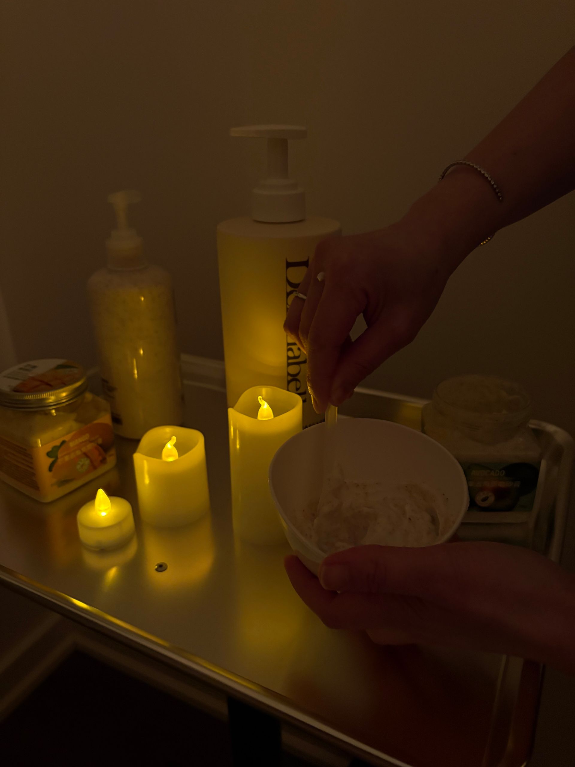 Hands placing a cotton pad near lit candles and skincare bottles in a dim bathroom