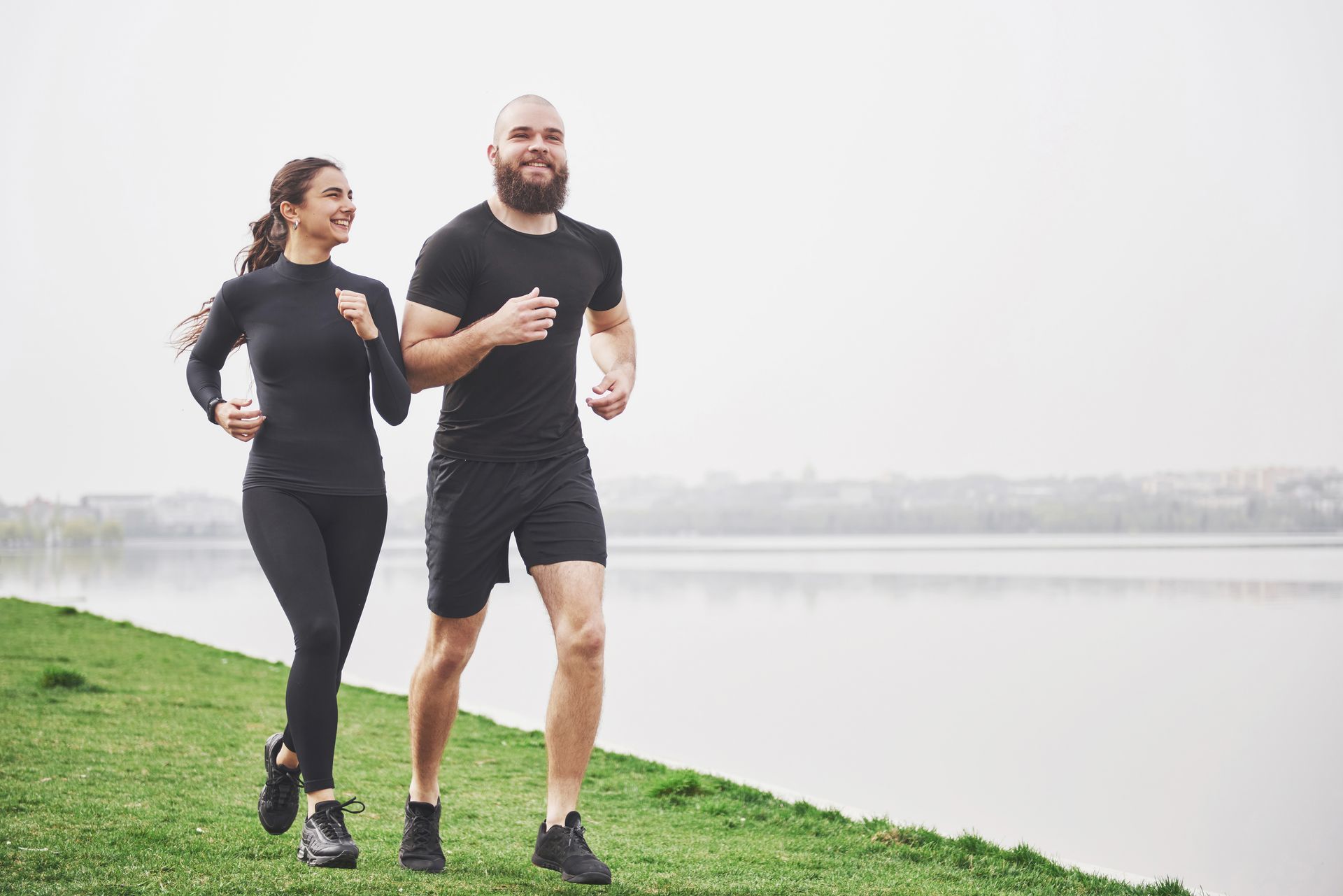 Two people jogging on a grassy path beside a calm lake on a foggy morning