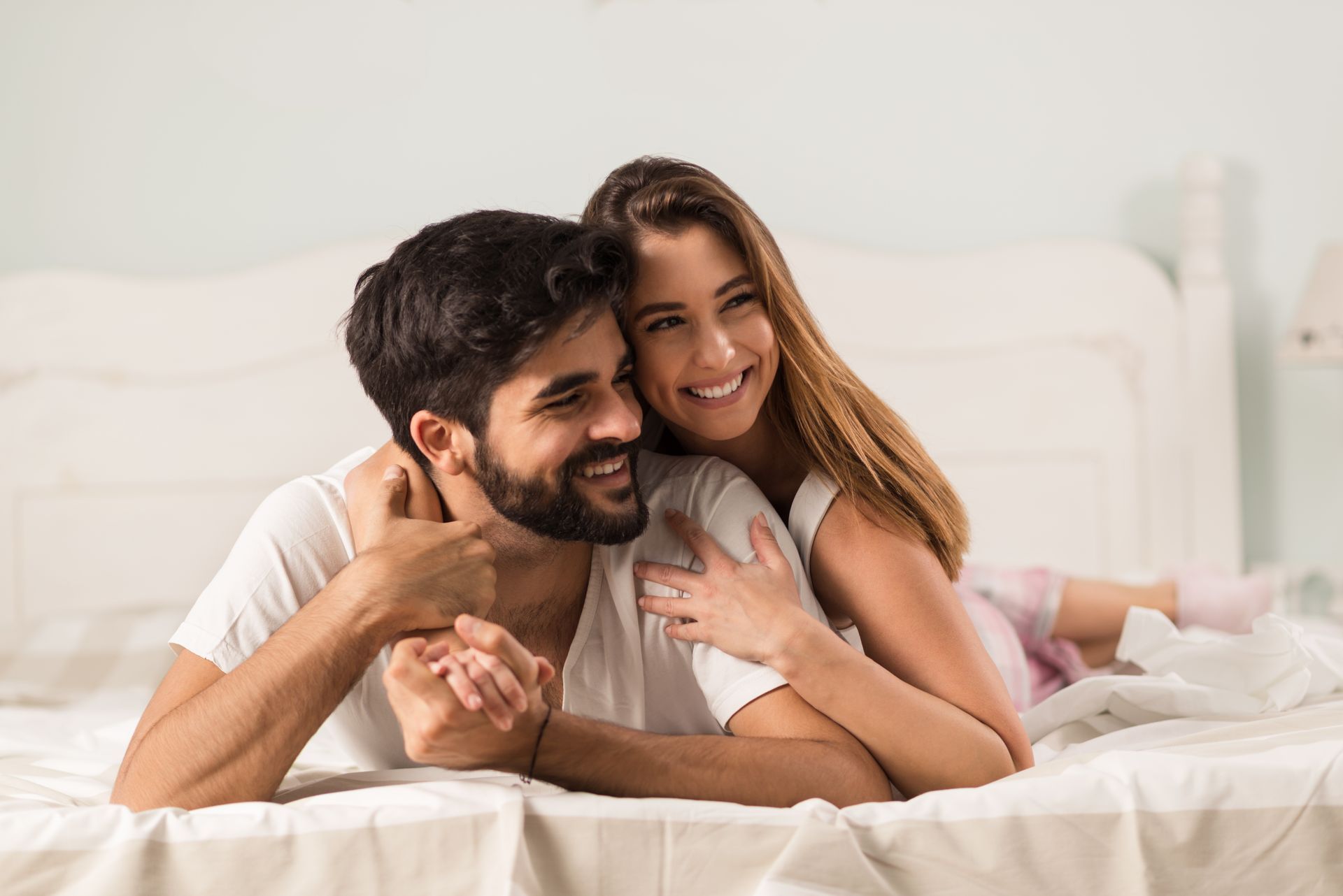 Couple smiling on a bed, embracing in a bright white bedroom
