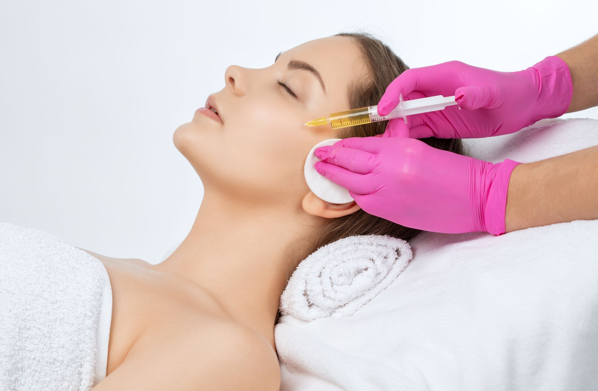 Person receiving facial injection; gloved hands injecting a clear substance into the cheek; medical setting.