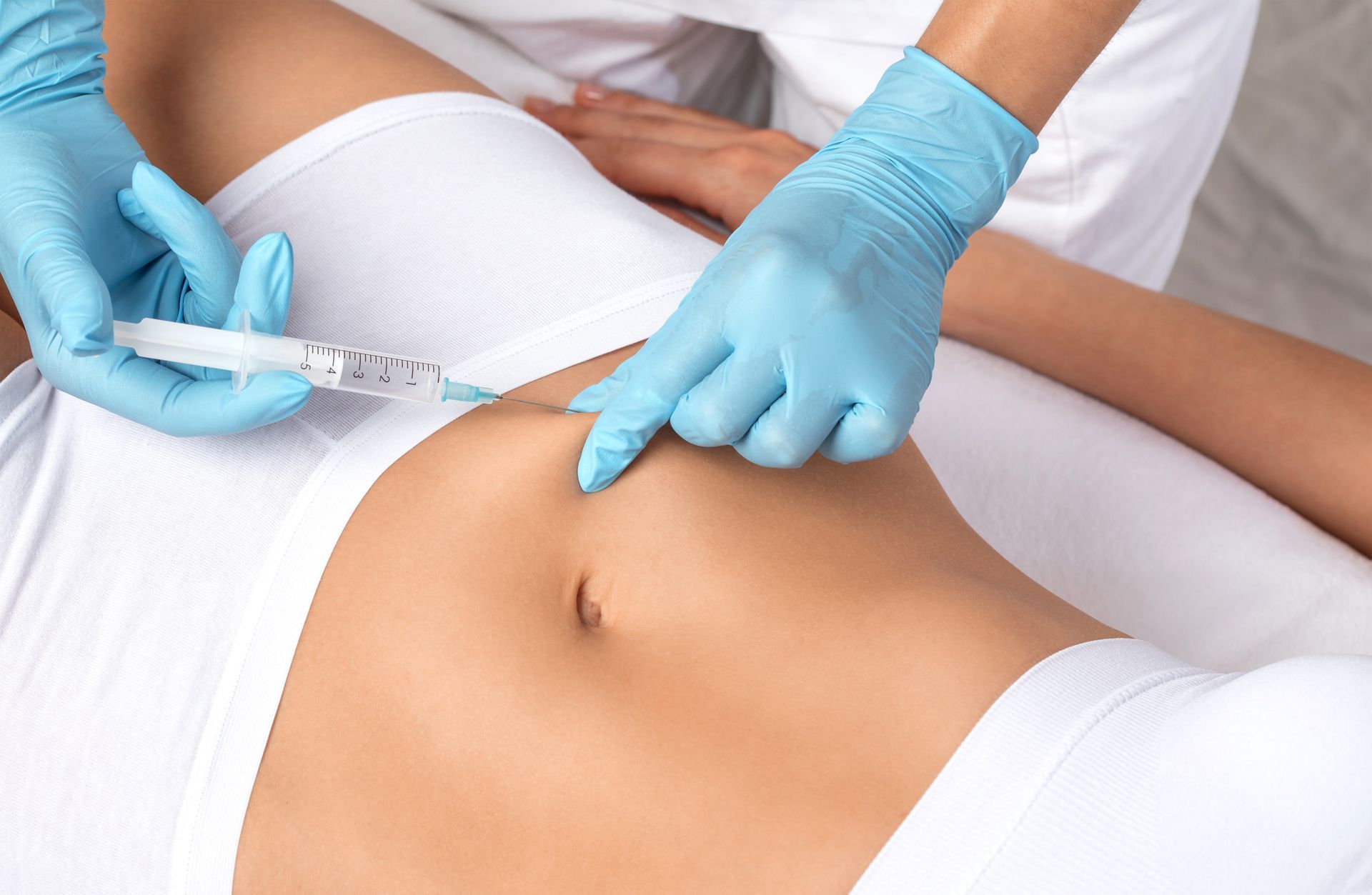 Gloved hands giving a syringe injection into a person’s abdomen on a medical bed