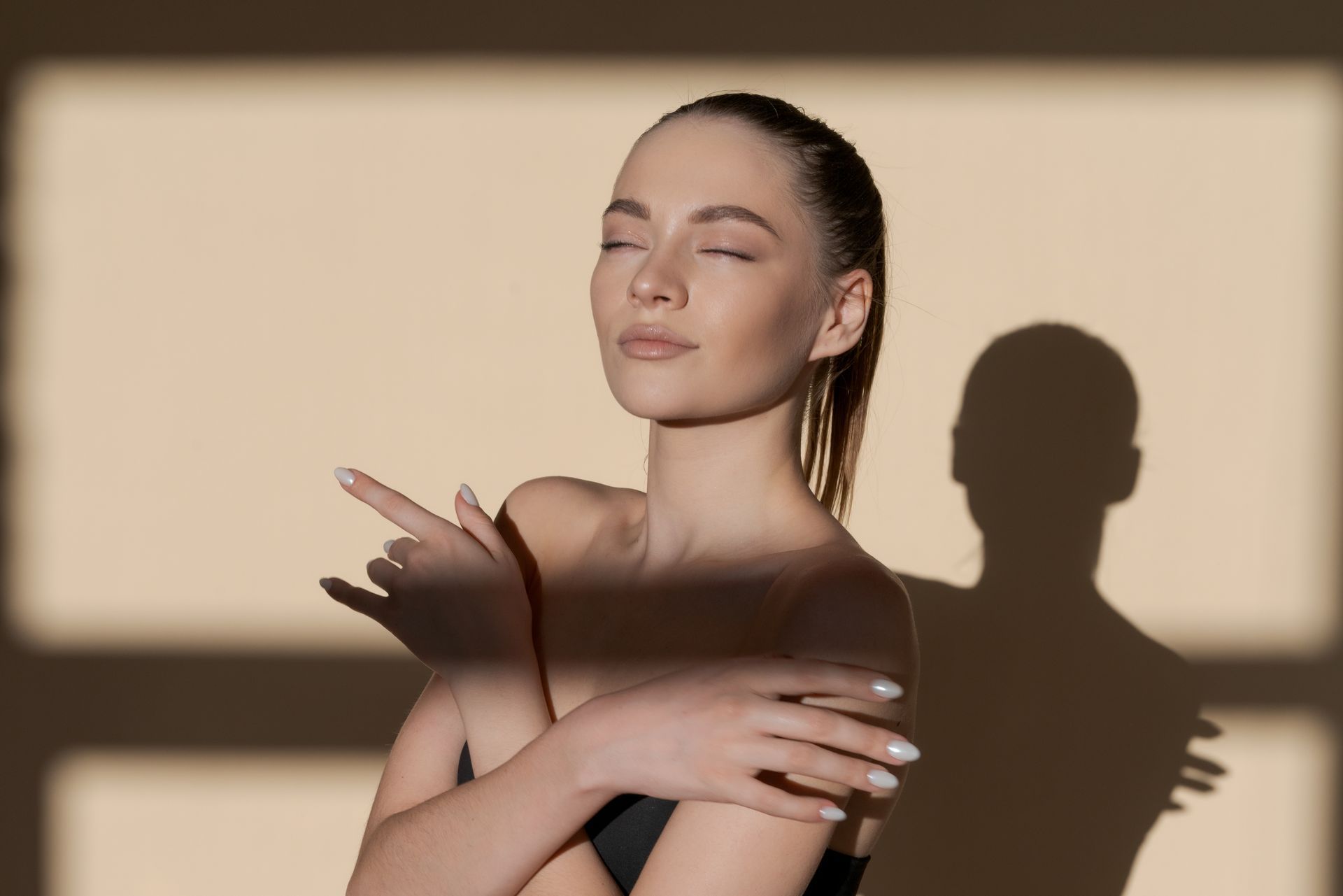 Woman with eyes closed posing in warm light, casting a shadow on a beige wall.