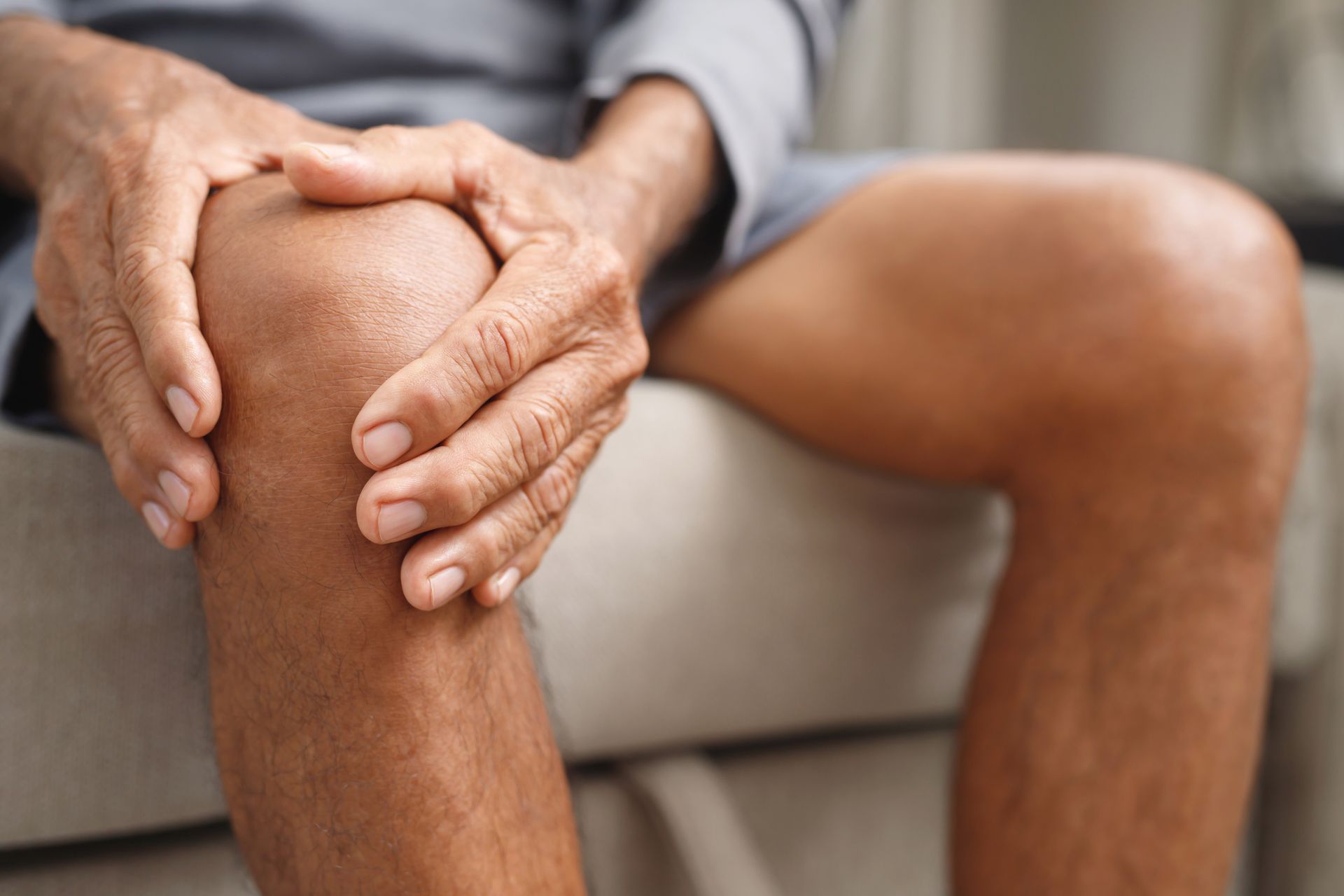 Person holding their knee, likely due to pain. Seated indoors. Dark skin, gray clothing.
