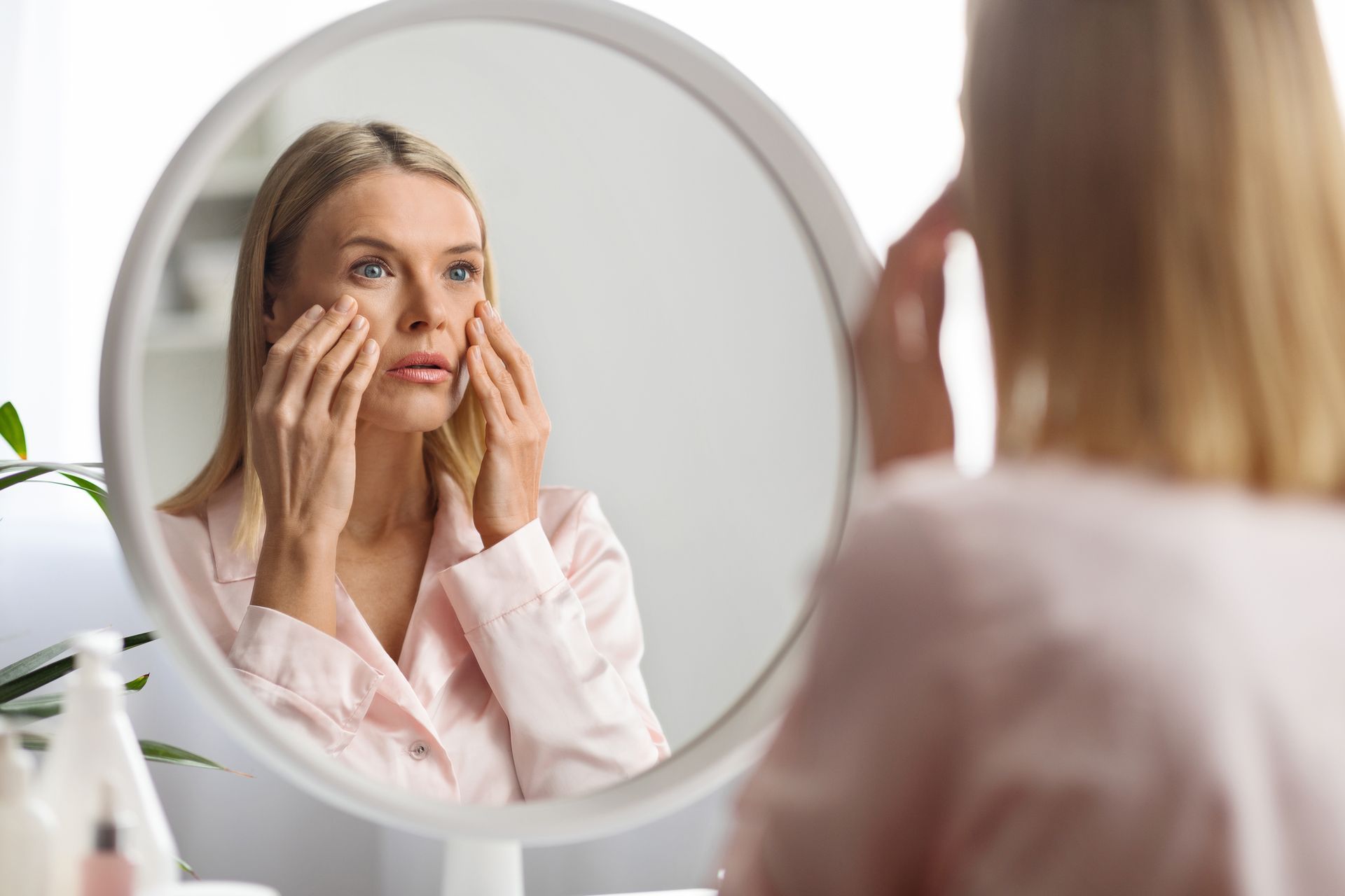 Woman in pink top looking in mirror, touching face, wrinkles visible.