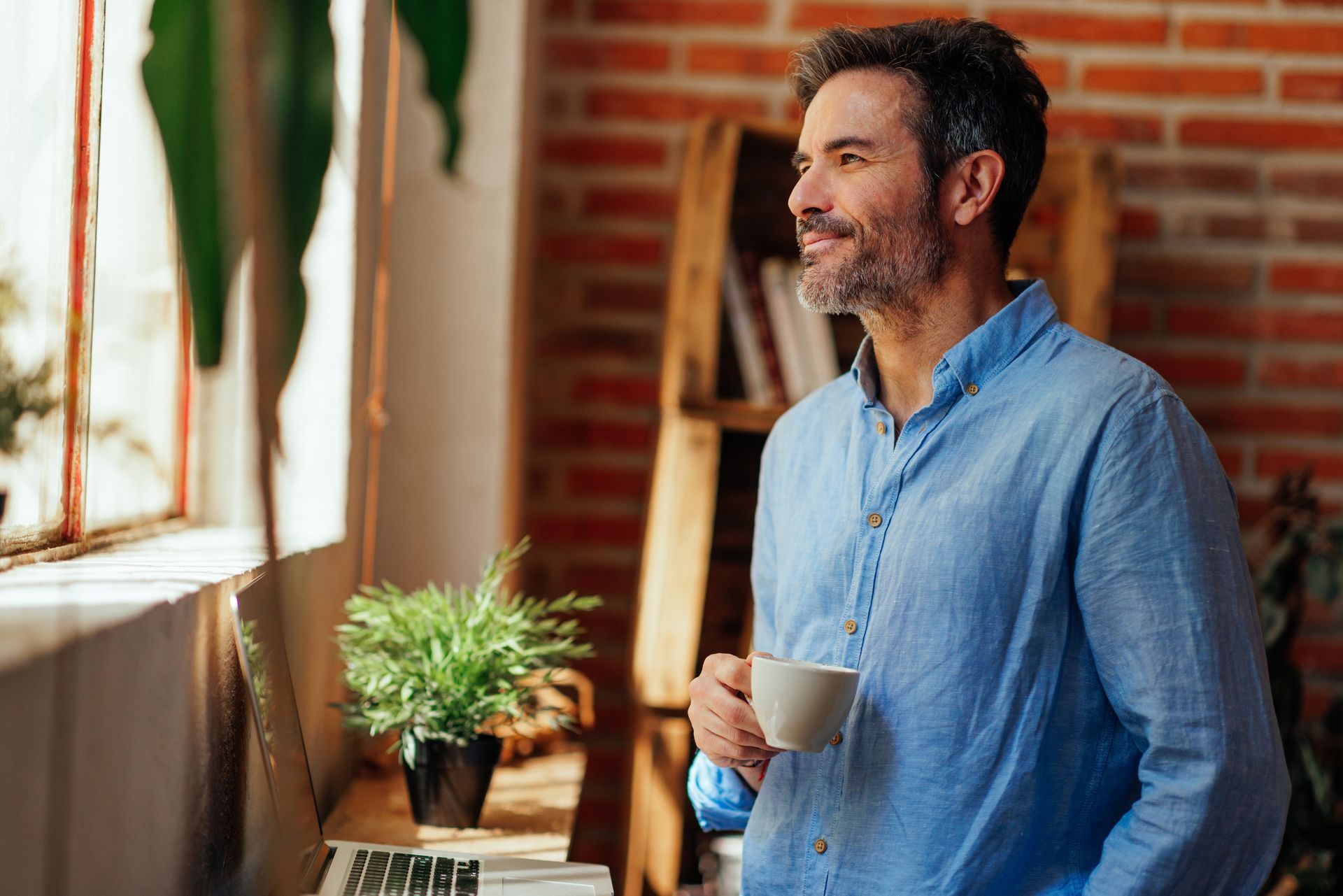 Man in blue shirt, holding mug, looking out window in a brick-walled room.