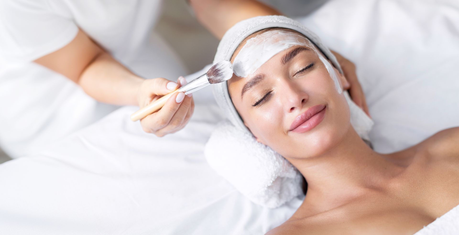 Person receiving a facial treatment while lying on a treatment bed, eyes closed, with white cream on the forehead.