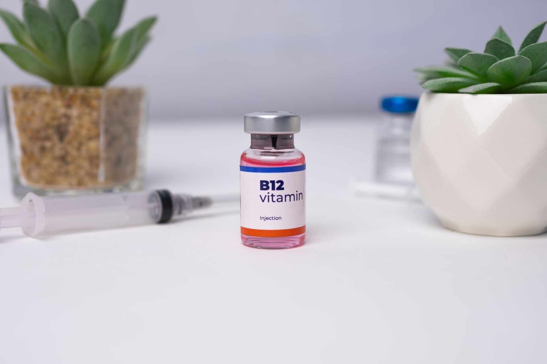 B12 vitamin bottle on a white table with a syringe and potted succulents in the background