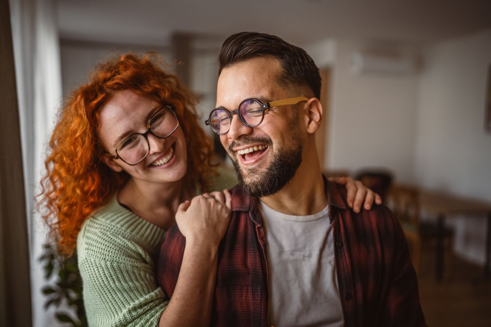 Woman with red curly hair and man with beard laughing, indoors.