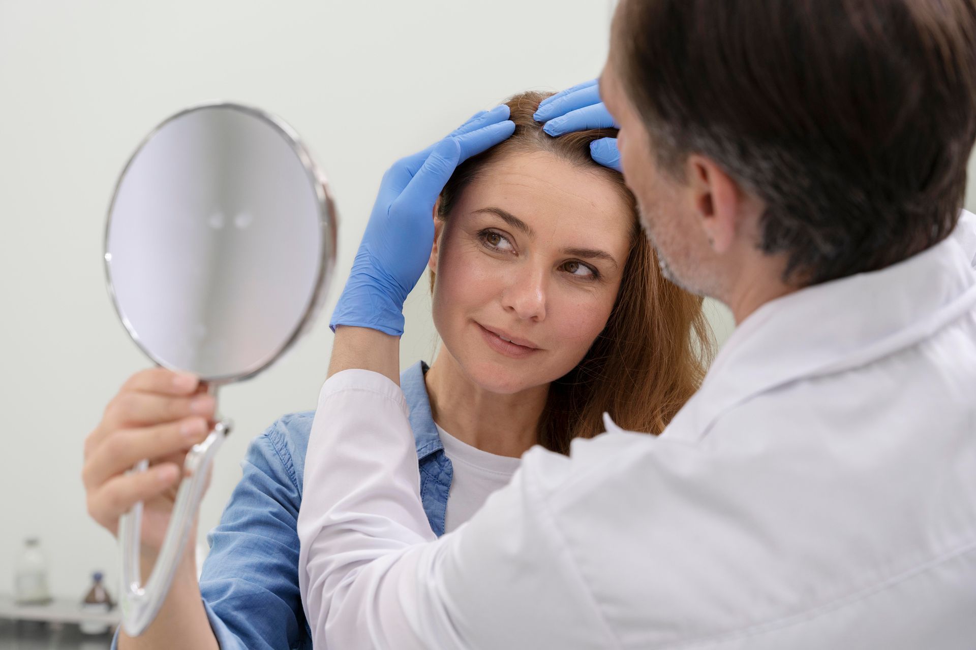 Doctor examining a patient’s forehead while a hand mirror reflects her face