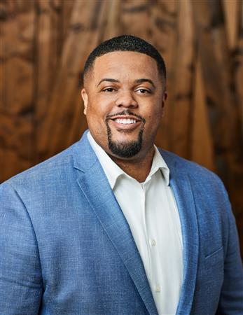 Man in blue blazer, smiling, against a wooden background.
