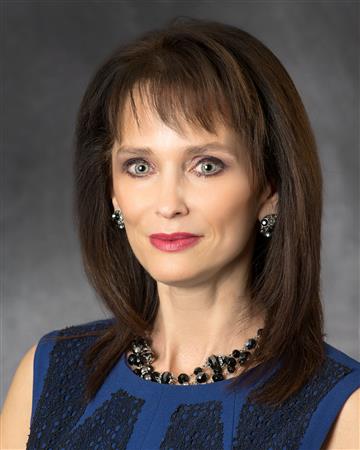 Woman in a blue dress and jewelry smiles, against a gray background.