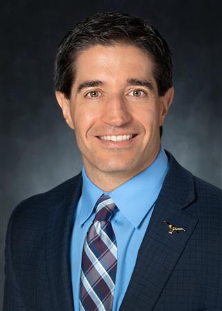 Man in suit smiling, light blue shirt, patterned tie. Dark background.