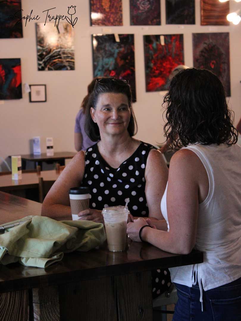 Two people chatting at a cafe table with coffee cups and wall art in the background.