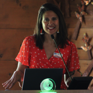 Woman in a red patterned blouse speaking at a podium with a laptop in a wooden room