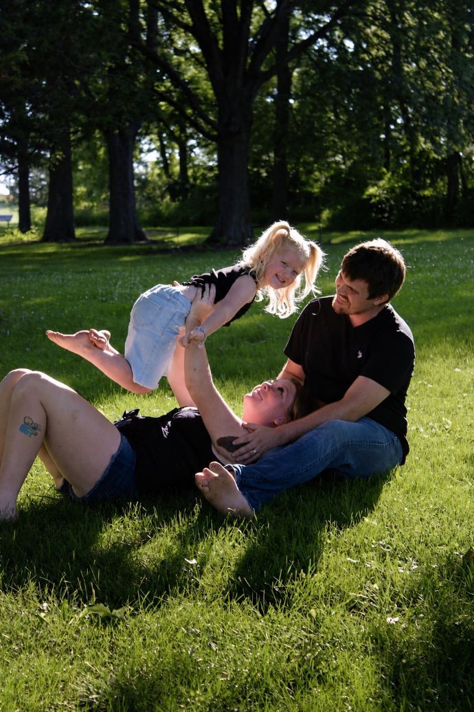 Two adults and a child play on grass in a sunny park, smiling and lying together.
