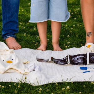 People standing barefoot on a white blanket with baby clothes and shoes on grass
