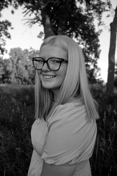 Black-and-white portrait of a smiling person with glasses outdoors near trees.