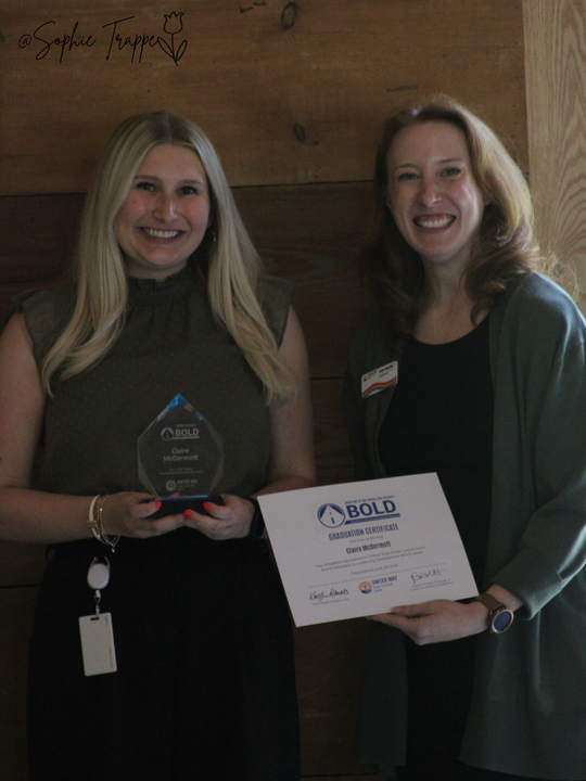 Two women smiling and holding an award plaque and certificate indoors