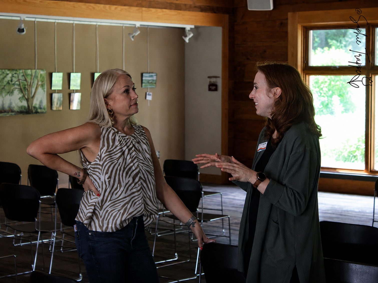 Two people talking in a wood-paneled room with chairs and a window.
