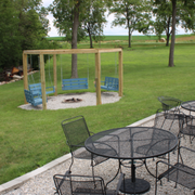 Backyard patio with black metal tables, chairs, and a fenced sand play area near a grassy field